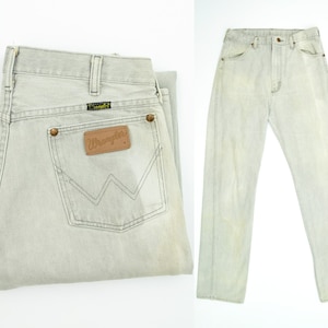 May include: A pair of light gray denim jeans with a brown leather Wrangler label on the back pocket. The jeans are folded in half and laid flat on a white surface.