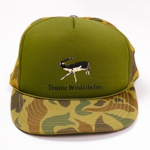 Peut inclure: Un chapeau de camionneur vert et camouflage avec une silhouette de cerf noir et blanc et le texte "Texotic Wildlife Inc."