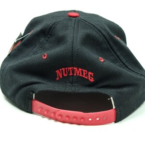Peut inclure: Casquette de baseball noire avec une sangle r&eacute;glable rouge et le mot "NUTMEG" brod&eacute; en rouge &agrave; l'arri&egrave;re.