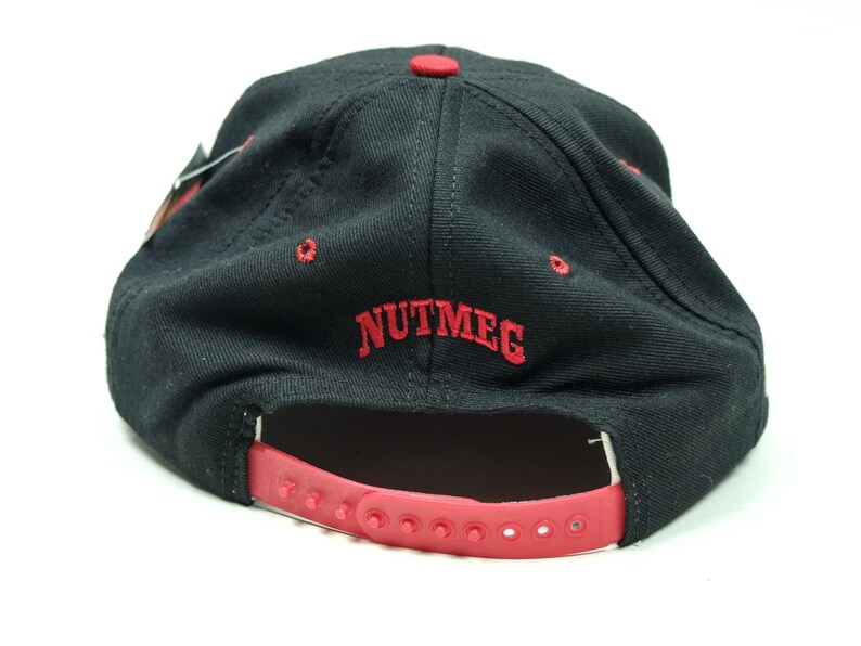 Peut inclure: Casquette de baseball noire avec une sangle r&eacute;glable rouge et le mot "NUTMEG" brod&eacute; en rouge &agrave; l'arri&egrave;re.