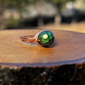 May include: A copper wire wrapped ring with a green iridescent gemstone. The ring is sitting on a wooden slice. The text "ROCK MY ZEN" is visible on the wooden slice.