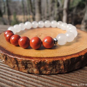 May include: A beaded bracelet featuring alternating red jasper and white quartz beads. The bracelet is strung on an elastic band and includes a small silver-colored spacer bead. The bracelet is displayed on a wooden slice.