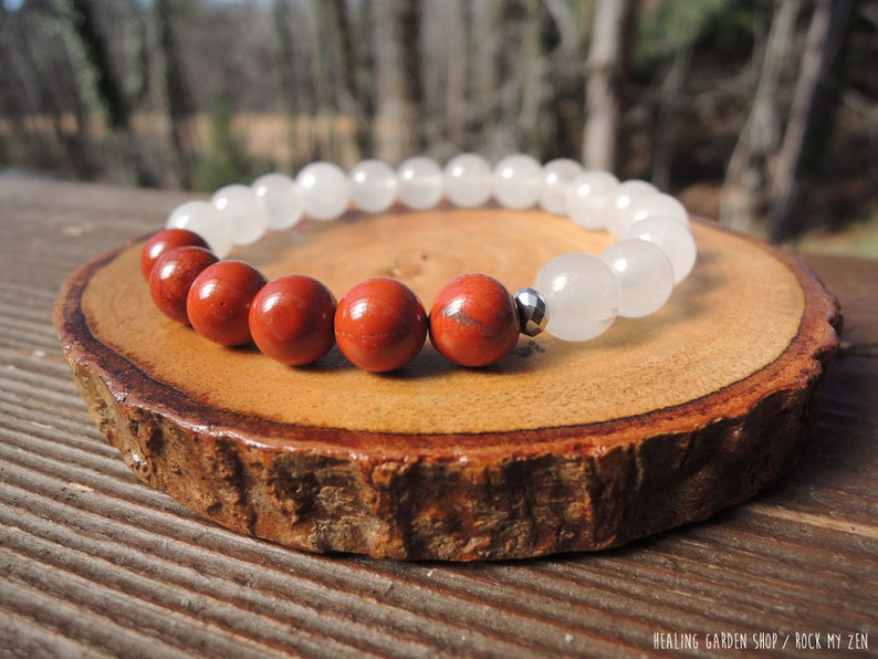 May include: A beaded bracelet featuring alternating red jasper and white quartz beads. The bracelet is strung on an elastic band and includes a small silver-colored spacer bead. The bracelet is displayed on a wooden slice.