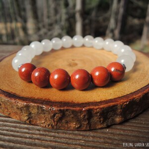 May include: A beaded bracelet featuring alternating white and red-brown gemstone beads. The bracelet is displayed on a round, wooden slice, with the text "HEALING GARDEN SHOP / ROCK MY ZEN" in the bottom right corner.
