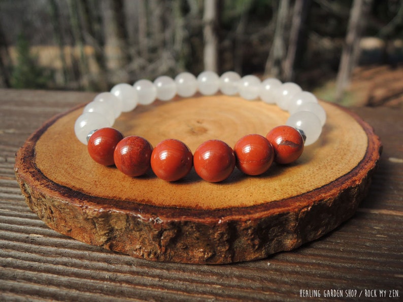 May include: A beaded bracelet featuring alternating white and red-brown gemstone beads. The bracelet is displayed on a round, wooden slice, with the text "HEALING GARDEN SHOP / ROCK MY ZEN" in the bottom right corner.