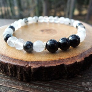 May include: A beaded bracelet featuring alternating clear and black beads. The bracelet is displayed on a round, wooden slice with a natural edge. The text "HEALING GARDENSHOP ROCKYZEN" is visible in the bottom right corner.