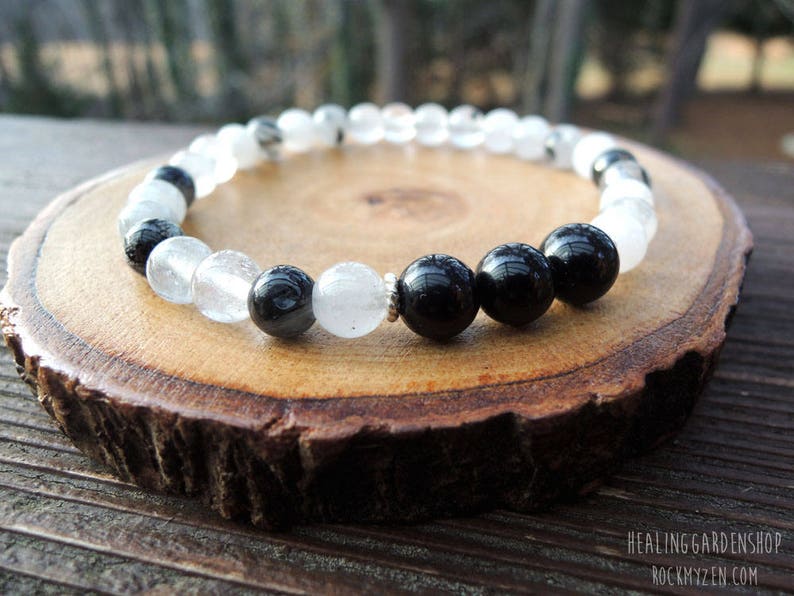 May include: A beaded bracelet featuring alternating clear and black beads. The bracelet is displayed on a round, wooden slice with a natural edge. The text "HEALING GARDENSHOP ROCKYZEN" is visible in the bottom right corner.