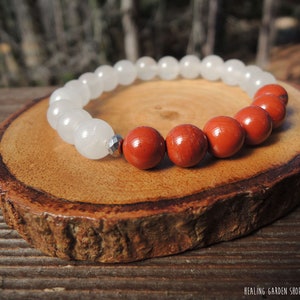 May include: A beaded bracelet featuring white and red-orange gemstone beads. The bracelet is displayed on a wooden slice. The text "HEALING GARDEN SHOP / ROCK MY ZEN" is visible in the bottom right corner.