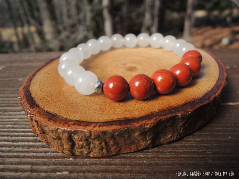 May include: A beaded bracelet featuring white and red-orange gemstone beads. The bracelet is displayed on a wooden slice. The text "HEALING GARDEN SHOP / ROCK MY ZEN" is visible in the bottom right corner.