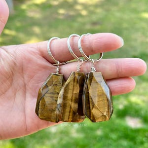May include: Three faceted tiger's eye keychains with silver-colored key rings. The gemstones have a warm brown color with natural patterns. The keychains are held in a hand against a blurred green background. The text "ROCK MY ZEN" is visible.