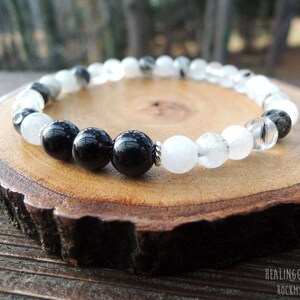 May include: A beaded bracelet featuring alternating black and white round beads. The bracelet is displayed on a wooden slice with a natural edge. The beads have a glossy finish, and the bracelet appears to be strung on a durable cord.