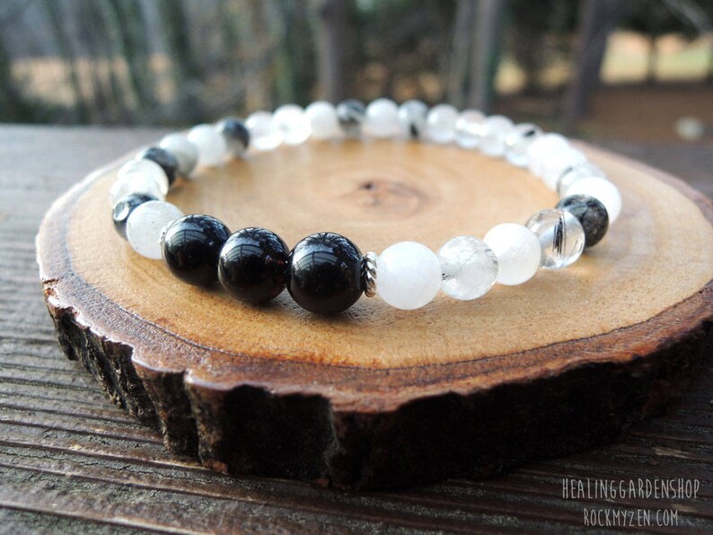 May include: A beaded bracelet featuring alternating black and white round beads. The bracelet is displayed on a wooden slice with a natural edge. The beads have a glossy finish, and the bracelet appears to be strung on a durable cord.