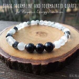 May include: A beaded bracelet featuring black tourmaline and tourmalated quartz. The bracelet has alternating black and clear beads, with the text "BLACK TOURMALINE AND TOURMALATED QUARTZ for Negative Energy Protection". The bracelet is displayed on a wooden slice.