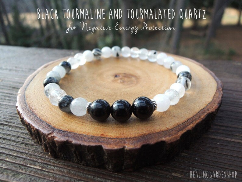 May include: A beaded bracelet featuring black tourmaline and tourmalated quartz. The bracelet has alternating black and clear beads, with the text "BLACK TOURMALINE AND TOURMALATED QUARTZ for Negative Energy Protection". The bracelet is displayed on a wooden slice.