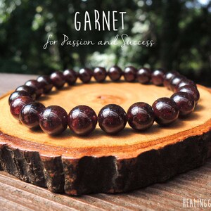 May include: A red garnet gemstone bracelet on a wooden surface. The bracelet is made of round, smooth beads. The text "GARNET for Passion and Success" is written above the bracelet.
