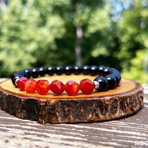 May include: A black and red gemstone bracelet with a silver bead accent. The bracelet is made with round black beads and faceted red beads. The bracelet is displayed on a wooden surface.