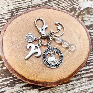 May include: Silver-toned keychain featuring various charms, including an Aries zodiac symbol, a crescent moon and star, and clear crystal beads. The keychain is displayed on a wooden slice with a natural wood grain.