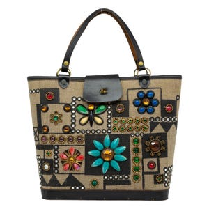 May include: A beige tote bag with a black leather handle and flap closure. The bag is decorated with colorful gemstones and a butterfly design. The bag is approximately 12 inches tall.