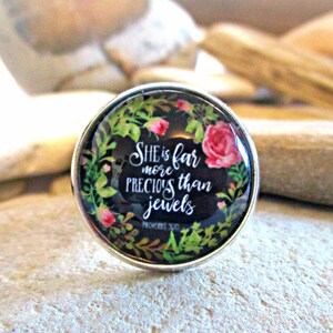 May include: A silver ring with a glass cabochon featuring a floral design and the text "She is far more precious than jewels Proverbs 31:10".