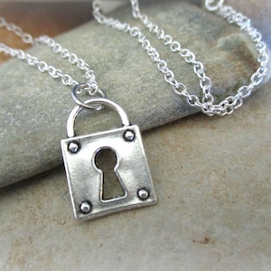 Puede incluir: Un collar de plata con un colgante de candado. El candado tiene una cerradura y está decorado con pequeños remaches.