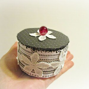 May include: A small, round decorative box with a gray lid and floral lace accents. The lid is adorned with a red gem and a white lace flower. The box is held in a hand.