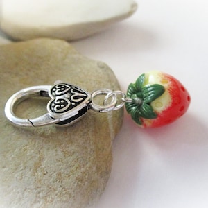 May include: A silver charm with a heart-shaped design and a red and white strawberry charm attached to it.