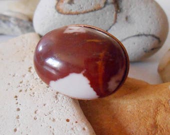 May include: A close-up of a red and white agate gemstone set in a copper ring. The gemstone has a smooth, polished surface and a swirling pattern of red and white.
