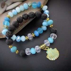 May include: Two bracelets with blue and clear glass beads, black lava rock beads, and gold accents. The bottom bracelet has a gold seashell charm.