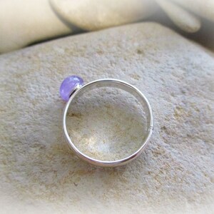 May include: A silver ring with a light purple gemstone. The ring is on a light brown surface.