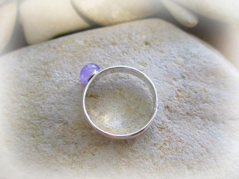 May include: A silver ring with a light purple gemstone. The ring is on a light brown surface.