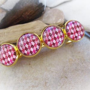 May include: Gold-toned hair barrette featuring four circular, pink and white checkered designs. The barrette is set against a neutral background of stones and wood.