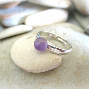May include: A silver ring with a round, light purple gemstone. The ring is sitting on a white rock.