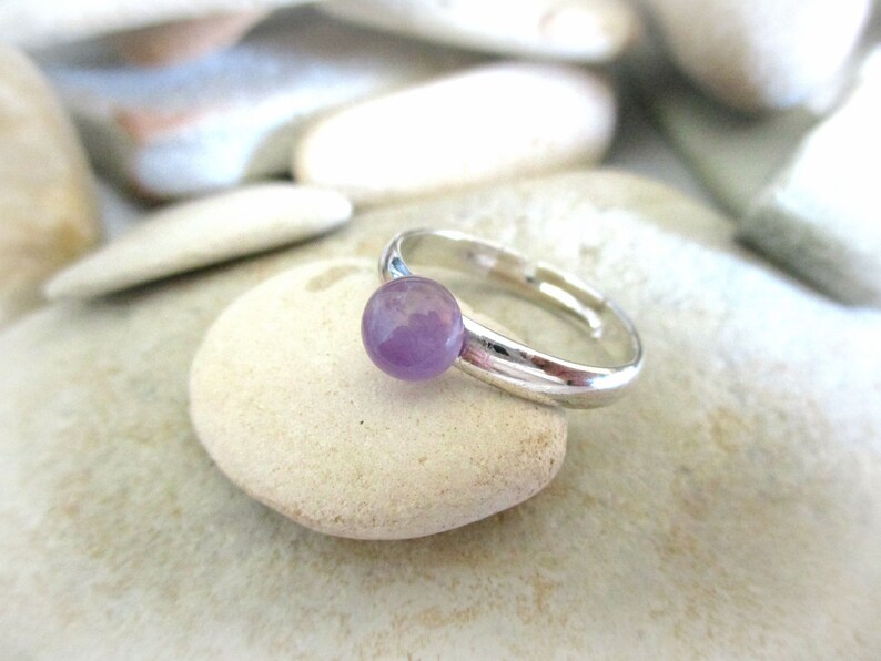 May include: A silver ring with a round, light purple gemstone. The ring is sitting on a white rock.