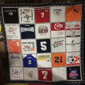 May include: A quilt made of various t-shirts, each with a different design and text. The quilt is made up of 16 squares, with each square containing a different t-shirt. The t-shirts are all different colors and have a variety of designs, including sports teams, school names, and other logos. The quilt is a great way to show off your love of sports and your school spirit.