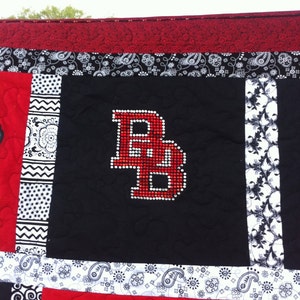 May include: A black and white quilt with a red letter "B" made of rhinestones in the center. The quilt is made of different fabrics with paisley and floral patterns.