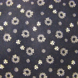 May include: A dark navy blue fabric featuring a repeating pattern of small white circular wreaths and tiny three-petaled floral motifs. The textile has a visible woven texture with a consistent, evenly spaced print across the entire surface area.
