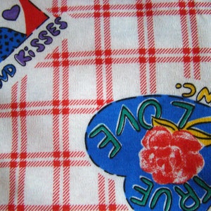 May include: A white fabric with a red and white plaid pattern. A blue heart with the words "TRUE LOVE" and "INC." is in the center of the fabric. The heart has a red rose inside and green leaves. The fabric has the words "HUGS AND KISSES" in the top left corner.