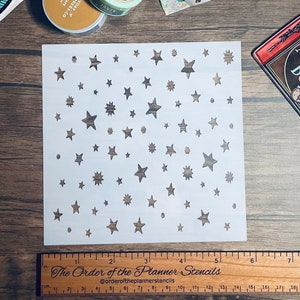 May include: A white plastic stencil with a repeating pattern of stars of different sizes. The stencil is 6 inches wide and 6 inches tall. The Order of the Planner Stencils is printed on a ruler below the stencil.