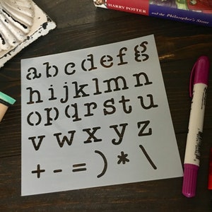 May include: A white stencil with black letters and symbols. The letters include the alphabet, plus signs, minus signs, an equals sign, a parenthesis, an asterisk, and a forward slash.