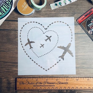 May include: A white stencil with a heart shape made of dotted lines. Inside the heart are four airplane stencils, with one airplane flying out of the heart. The stencil is 15.2 cm wide and 15.2 cm tall.