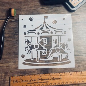 May include: White plastic stencil of a carousel with three horses, a roof, and stars. The stencil is 6 inches wide and 6 inches tall. The Order of the Planner Stencils logo is printed on a wooden ruler below the stencil.
