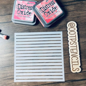 May include: White plastic stencil with 12 horizontal lines. The stencil is on a wooden surface with two ink pads labeled "Distress Oxide" in pink lettering. The ink pads are labeled "picked raspberry" and "festive berries".
