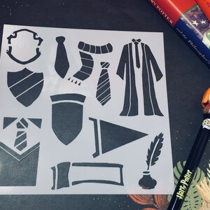 May include: A white stencil with black cutouts of various Harry Potter themed images, including a tie, a scarf, a robe, a crest, and a quill. The stencil is on a black surface.
