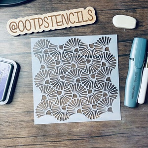 May include: A white plastic stencil with a repeating pattern of overlapping fan shapes. The stencil is on a wooden surface with a wooden sign that says "@OOTPSTENCILS".