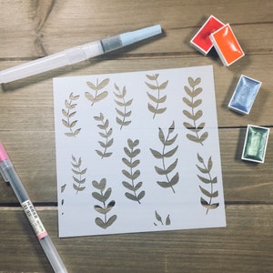 May include: A white stencil with a design of various leaf shapes. The stencil is on a wooden surface with watercolor paints and a paintbrush.