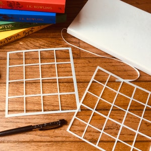 May include: Two white plastic stencils with a grid pattern. Each stencil has 36 squares. A black pen is lying on a wooden surface.