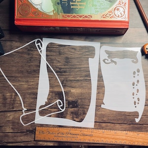 May include: Three white stencils for crafting. The stencils are shaped like a rolled up scroll, a boot, and a boot print. The stencils are on a wooden surface with a ruler.