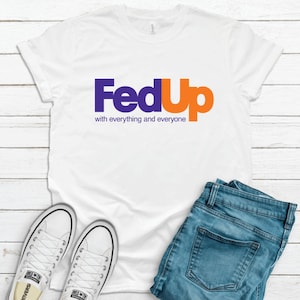 May include: White t-shirt with a purple and orange logo that says "FedUp with everything and everyone". The shirt is paired with blue jeans and white sneakers.