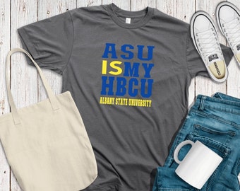 May include: A gray t-shirt with the text "ASU IS MY HBCU" in blue and yellow letters. The text "ALBANY STATE UNIVERSITY" is printed in smaller letters below the main text. The shirt is laid out on a white wooden surface with a white tote bag, a pair of white sneakers, and a white mug.
