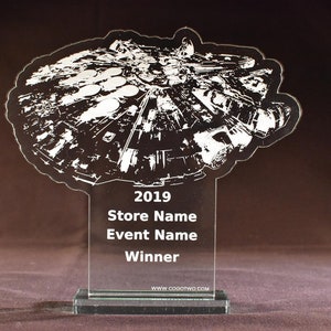 Peut inclure: Un trophée en verre transparent avec une silhouette blanche du vaisseau spatial Millennium Falcon. Le trophée porte les inscriptions "2019", "Nom du magasin", "Nom de l'événement" et "Gagnant" sur le socle. Le bas du socle porte l'inscription "www.cog0two.com".